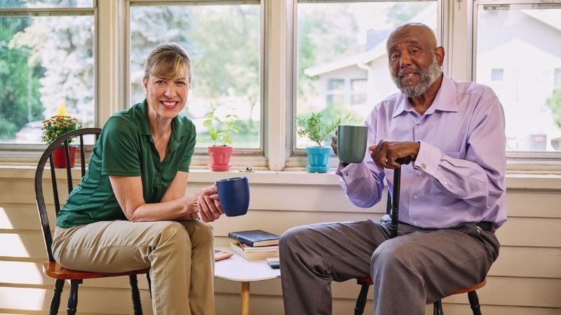 A confident Home Instead caregiver sitting with a client in the kitchen with coffe mugs