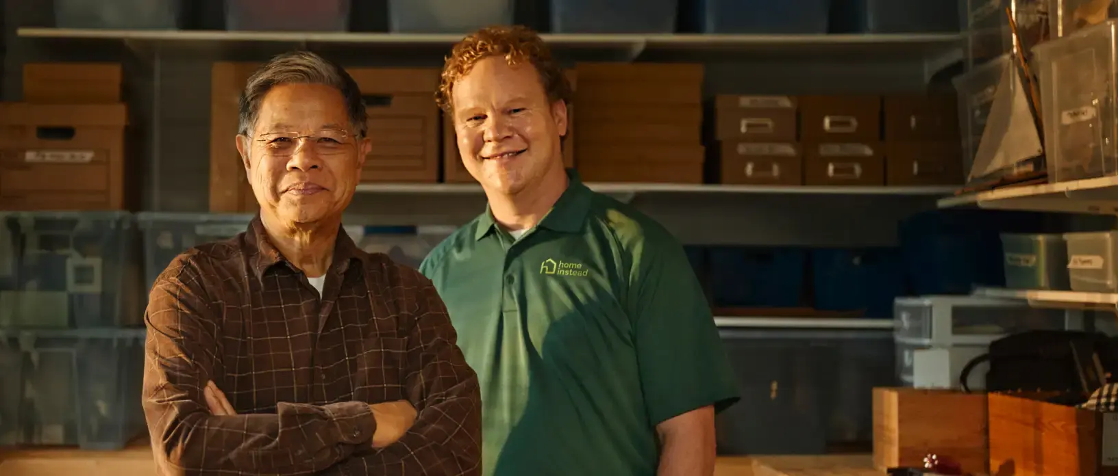 A Home Instead  caregiver in a green uniform stands next to an elderly man in a workspace, both smiling, symbolizing trust and collaboration.