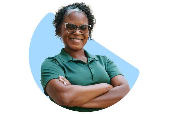 A confident Home Instead caregiver in a green uniform, smiling with arms crossed. The image has a circular blue background crop, emphasizing her approachable demeanor.