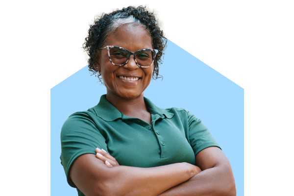 A confident Home Instead caregiver in a green uniform smiles with arms crossed, standing outdoors, representing trust and professionalism.