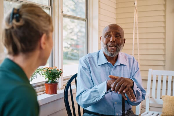 An elderly man gestures while speaking with his Home Instead caregiver on a bright porch, fostering a moment of friendly conversation and companionship.