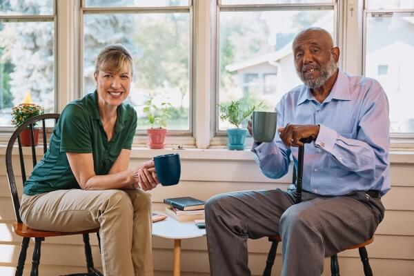 A Home Instead caregiver and an elderly man enjoy coffee together on a porch, showcasing shared moments of relaxation and connection.