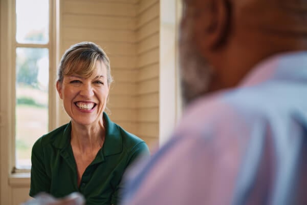 A Home Instead caregiver attentively listens to an elderly man as they chat in a sunlit room, highlighting meaningful connection and understanding.