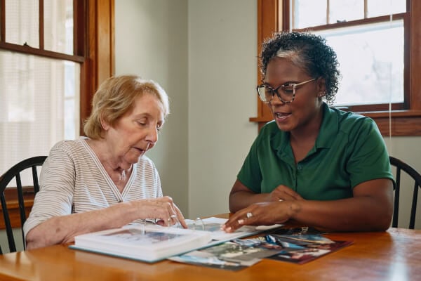 A Home Instead caregiver and an elderly woman sit together at a table, flipping through a scrapbook, highlighting moments of reminiscence and connection.