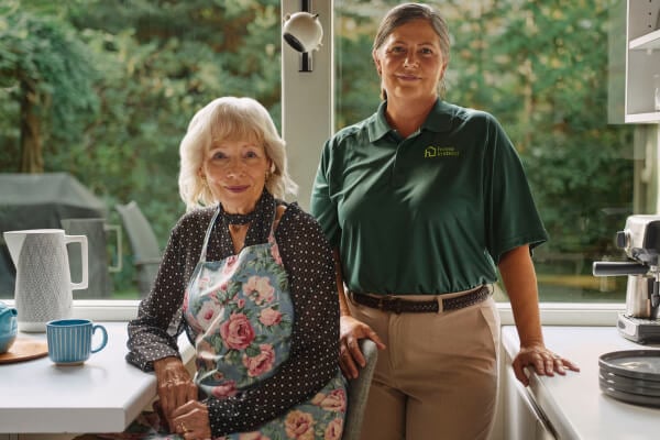 A Home Instead caregiver in a green uniform stands beside an elderly woman in a floral apron in a bright kitchen, symbolizing meal prep, support and collaboration.