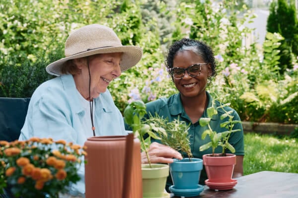 A cheerful elderly woman wearing a sun hat gardens alongside her Home Instead caregiver in a colorful and vibrant outdoor setting.