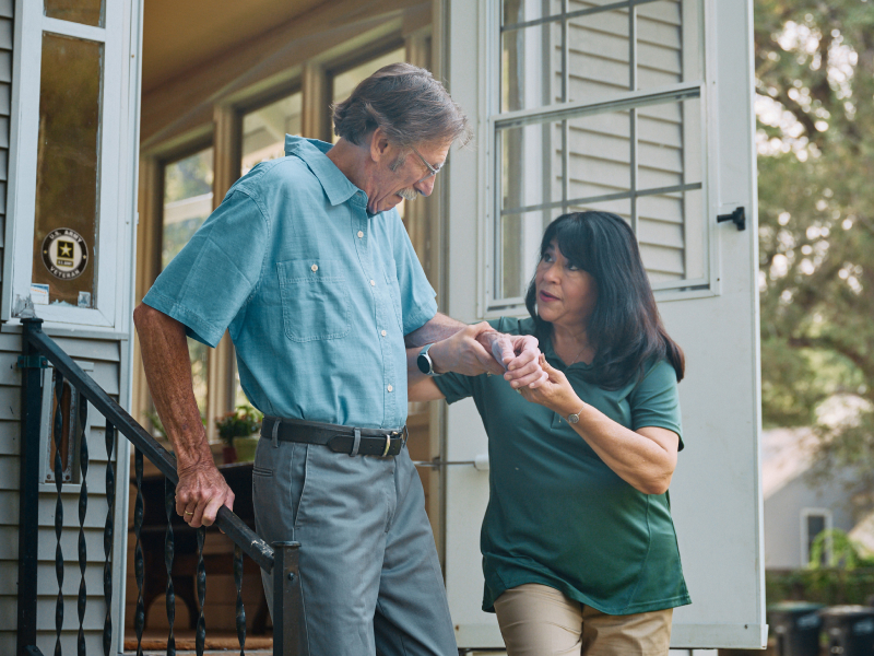 A Home Instead caregiver helps an elderly man, a veteran, safely walk down the front steps of a home, emphasizing Home Instead's care and thoughtful support.