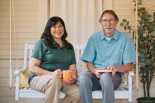 A Home Instead caregiver and an elderly man sit together on a porch swing, enjoying coffee and a book, emphasizing relaxed and meaningful companionship.