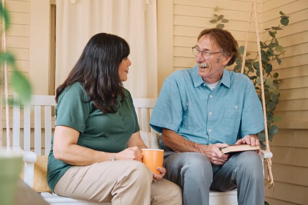 A Home Instead caregiver sits on a porch swing with an elderly man, both smiling warmly, highlighting a moment of shared connection and companionship.