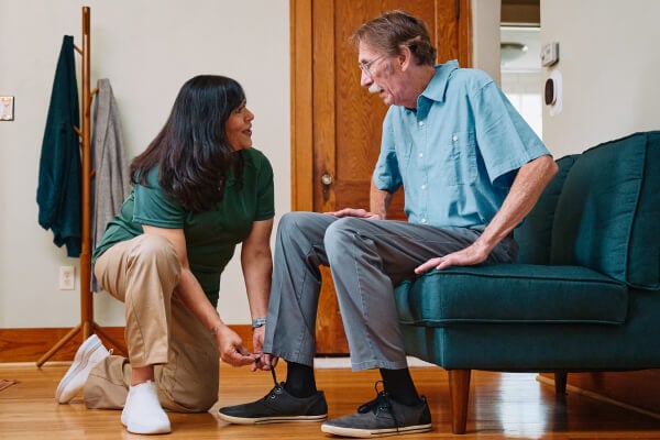 A Home Instead caregiver kneels to help an elderly man tie his shoes in a cozy living room, highlighting patient and personalized care for daily tasks.