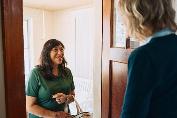 A Home Instead caregiver greets an elderly woman at the door with a warm smile, holding a tote bag, symbolizing friendliness and readiness to assist.