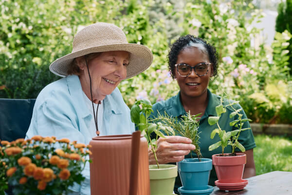 A Home Instead caregiver and an elderly woman smile together as they tend to potted plants outdoors, emphasizing companionship and shared hobbies.