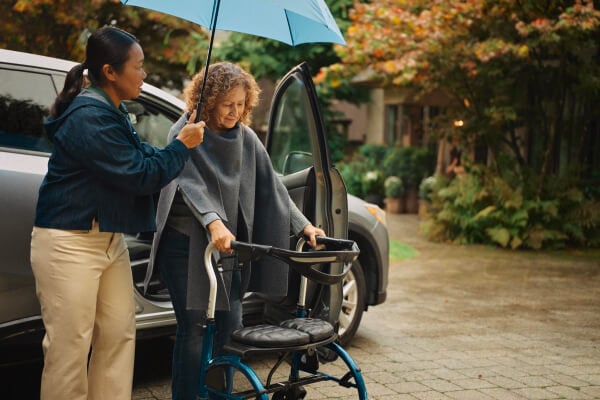 A Home Instead caregiver holds an umbrella for an elderly woman using a walker as they step out of a car on a rainy day, highlighting attentive and thoughtful care.