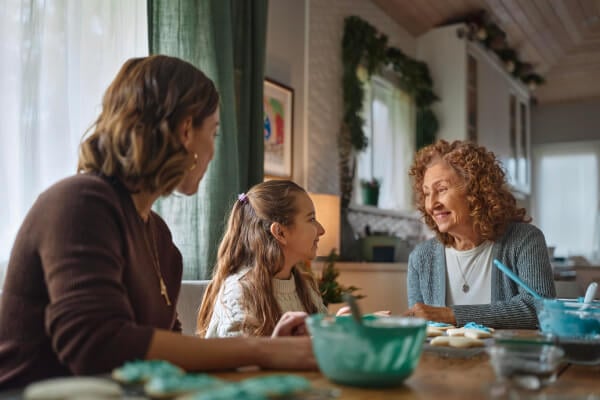 Three generations gathered around a table: an elderly woman, her daughter, and granddaughter baking cookies together, creating joyful family moments.