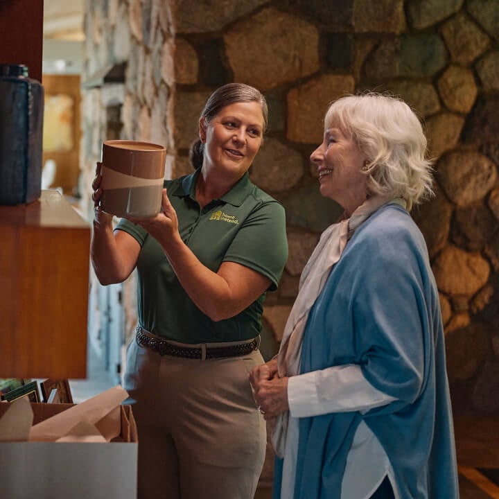 A Home Instead caregiver in a green uniform helps an elderly woman examine a piece of pottery in a warmly lit home, symbolizing companionship and support with daily activities.