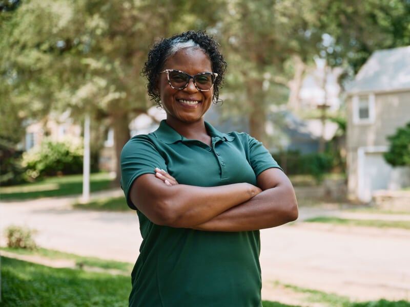 A confident Home Instead caregiver stands outdoors with arms crossed, smiling warmly, symbolizing professionalism and approachability.