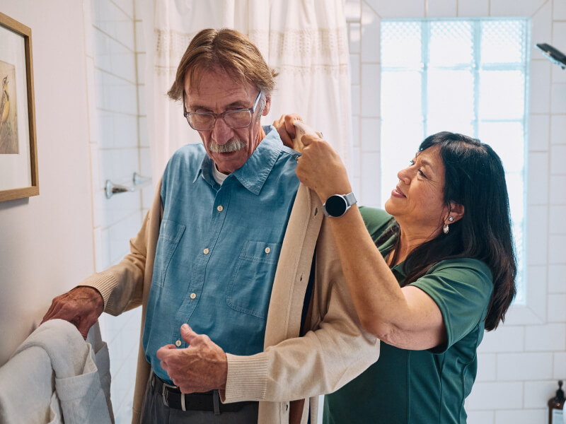 A Home Instead caregiver helps an elderly man adjust his sweater in a bathroom, emphasizing dignity and personalized care during daily routines.