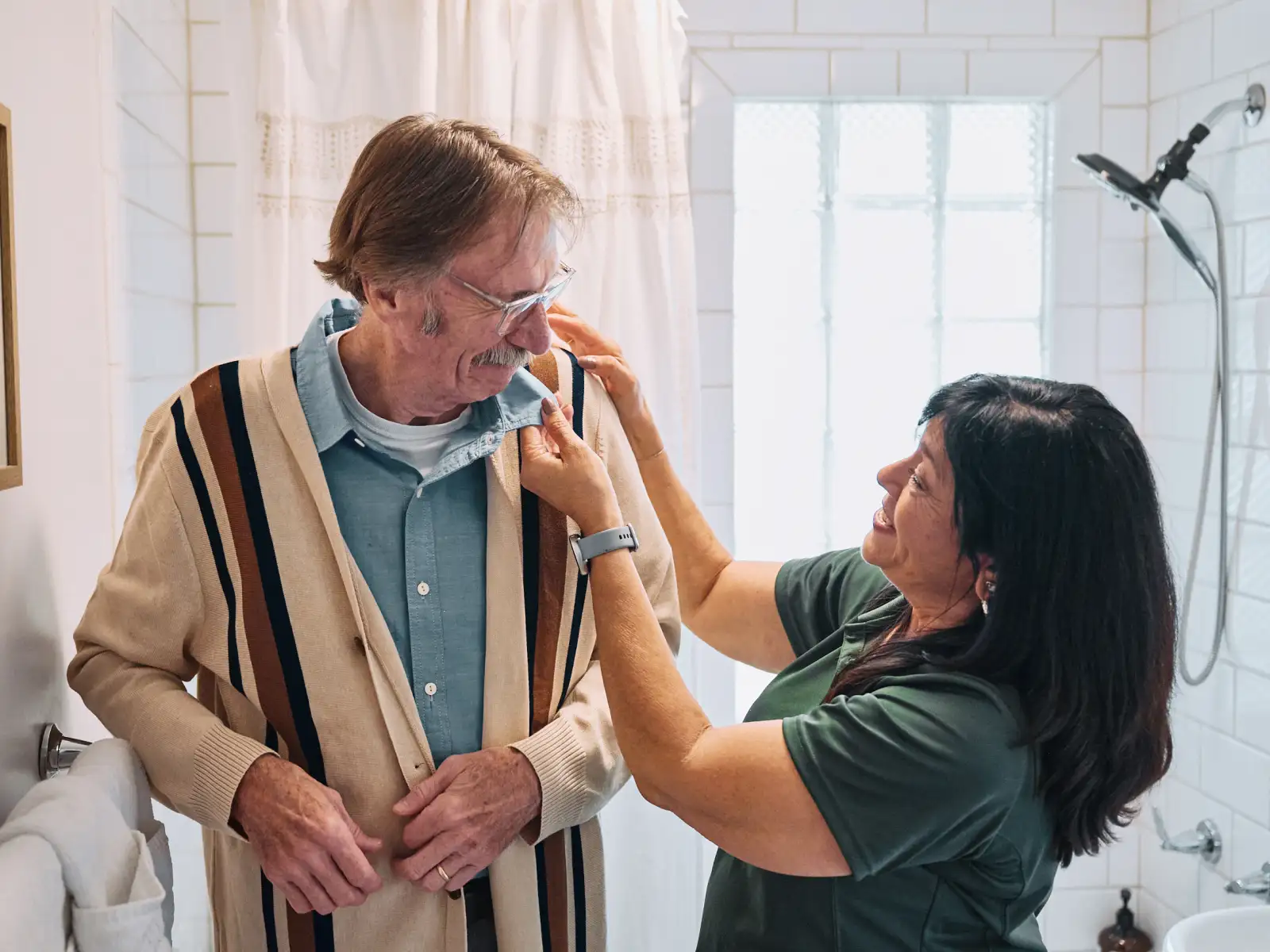 A Home Instead caregiver assists an elderly man with a cardigan sweater in a well-lit bathroom, emphasizing compassionate support for daily activities.