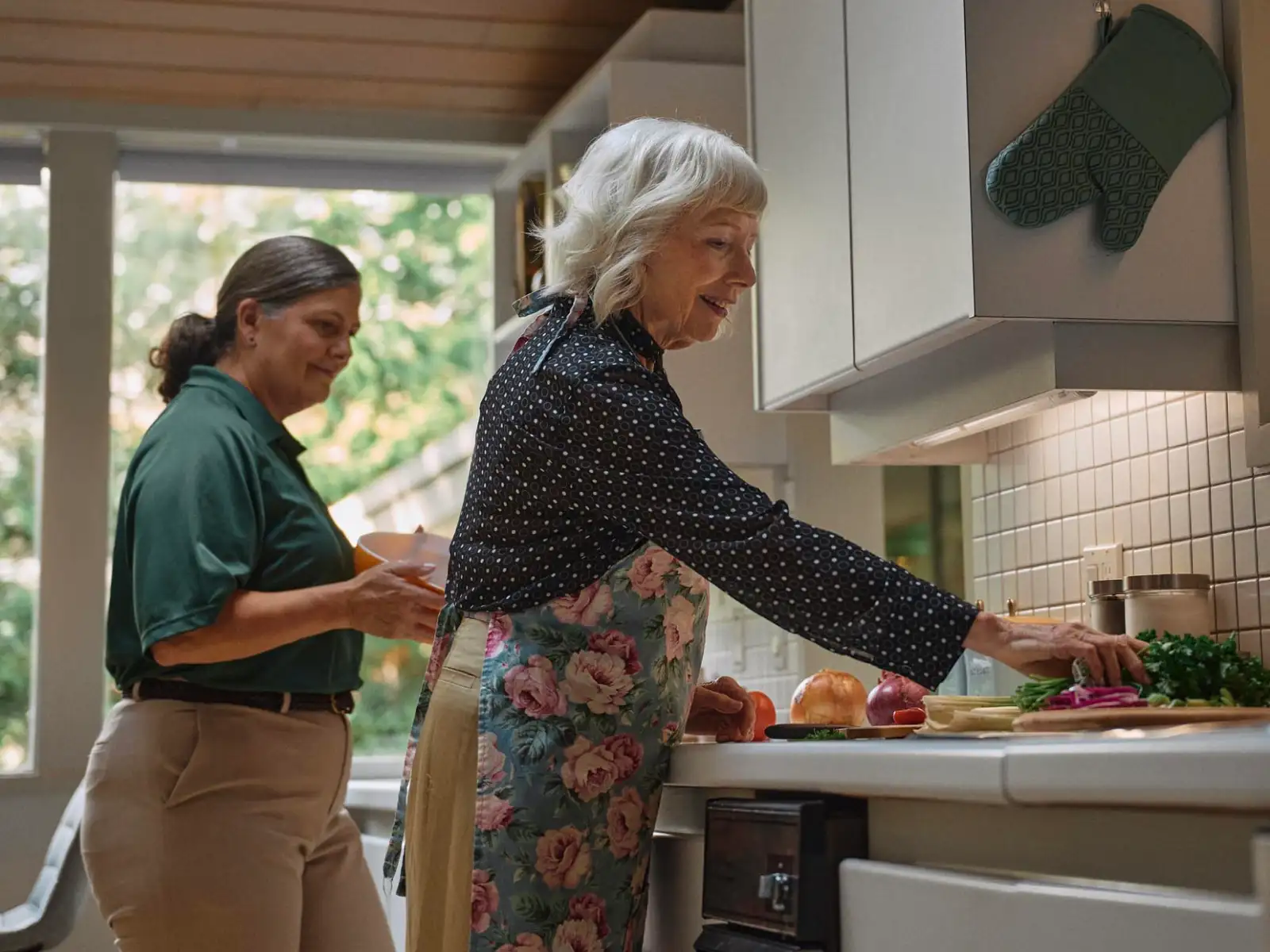 A Home Instead caregiver and an elderly woman prepare a meal together in a sunny kitchen, symbolizing teamwork and independence in home care.