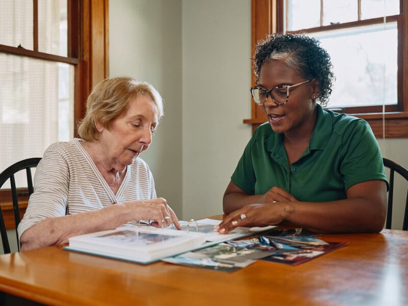 A Home Instead caregiver and an elderly woman sit together at a table, flipping through a scrapbook, highlighting moments of reminiscence and connection.