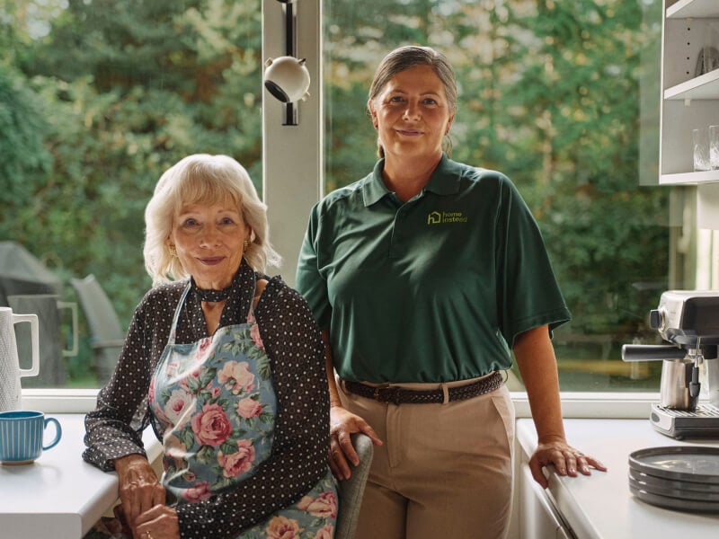 A Home Instead caregiver in a green uniform stands beside an elderly woman in a floral apron in a bright kitchen, symbolizing meal prep, support and collaboration.