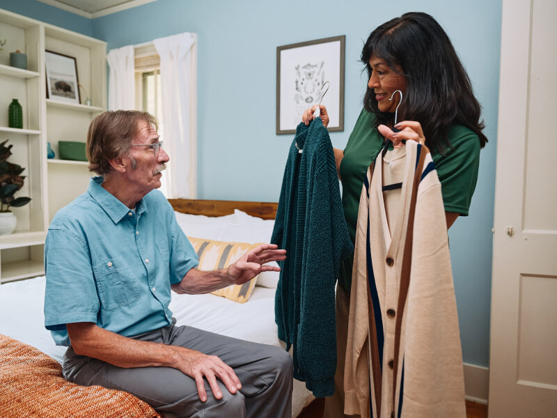 A Home Instead caregiver helps an elderly man choose between two sweaters, showcasing thoughtful and personalized assistance for daily routines.