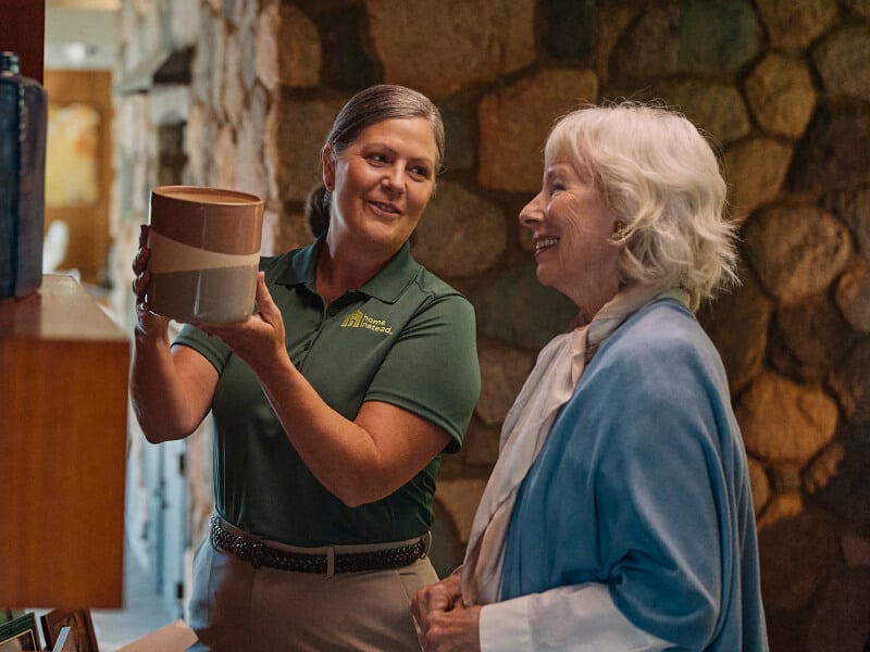 A Home Instead caregiver in a green uniform helps an elderly woman examine a piece of pottery in a warmly lit home, symbolizing companionship and support with daily activities.