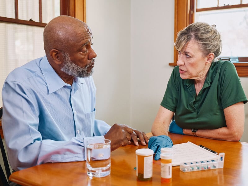 A Home Instead caregiver and an elderly man sit at a table discussing medications, with pill organizers and water, showcasing careful health management.