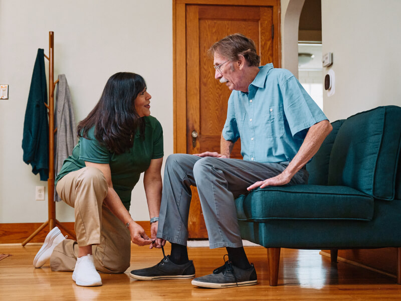 A Home Instead caregiver kneels to help an elderly man tie his shoes in a cozy living room, highlighting patient and personalized care for daily tasks.