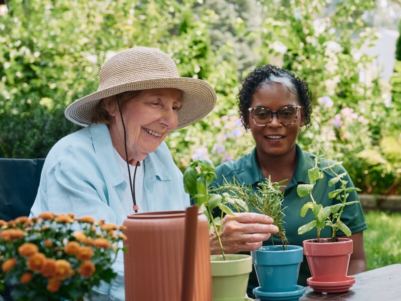 A Home Instead caregiver and an elderly woman smile together as they tend to potted plants outdoors, emphasizing companionship and shared hobbies.