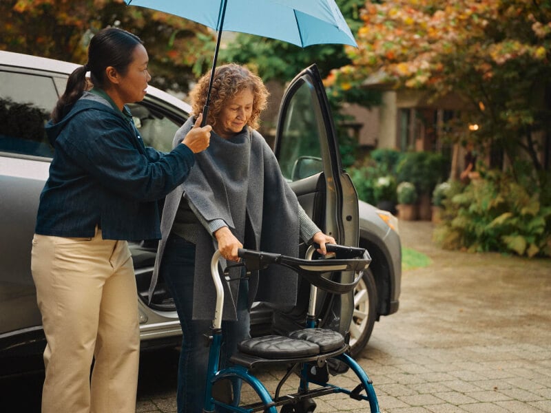 A Home Instead caregiver holds an umbrella for an elderly woman using a walker as they step out of a car on a rainy day, highlighting attentive and thoughtful care.