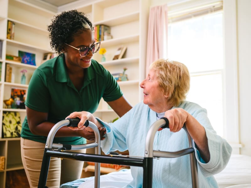 A smiling Home Instead caregiver in a green uniform assists an elderly woman using a walker in a bright and welcoming room, emphasizing support and encouragement.