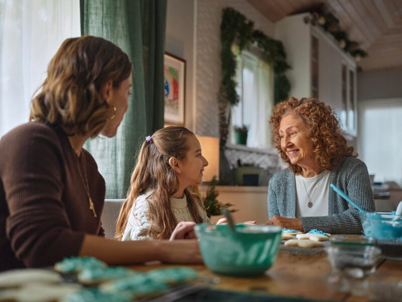 Three generations gathered around a table: an elderly woman, her daughter, and granddaughter baking cookies together, creating joyful family moments.