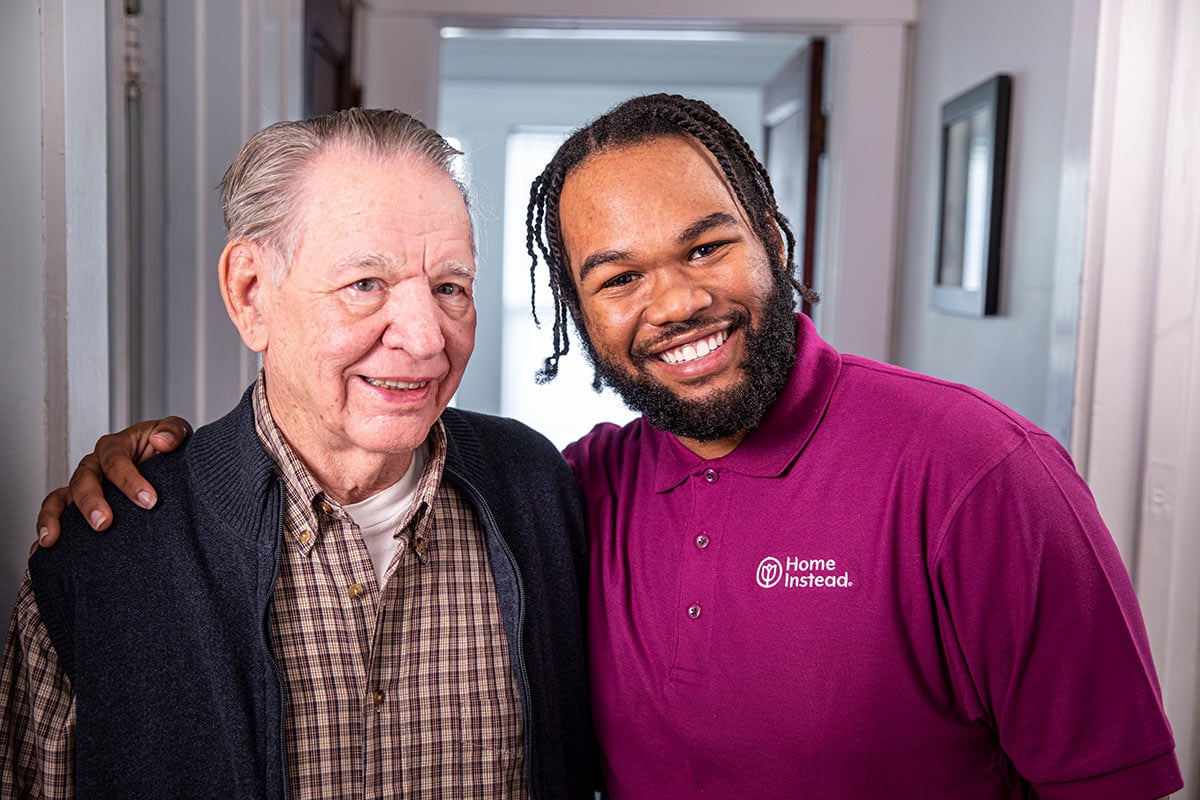 Home Instead Caregiver and senior man stand smiling together in home