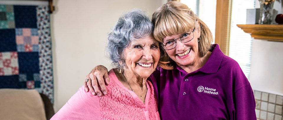 Home Instead Caregiver and senior woman with cane stand smiling together in living room